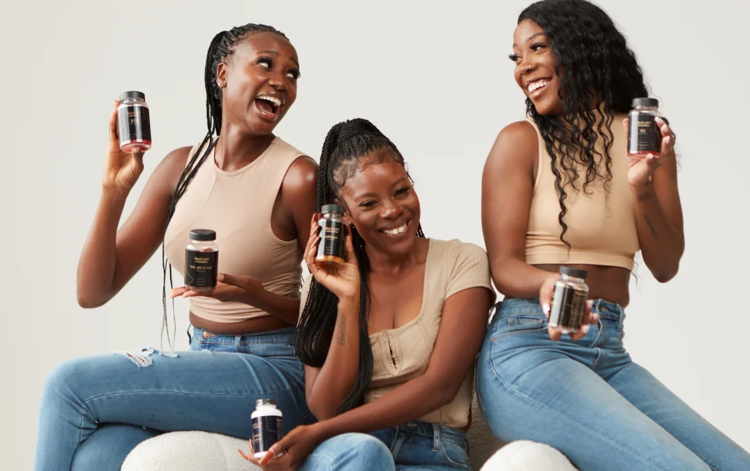 Three women laughing together on a couch, each holding a bottle of vitamins